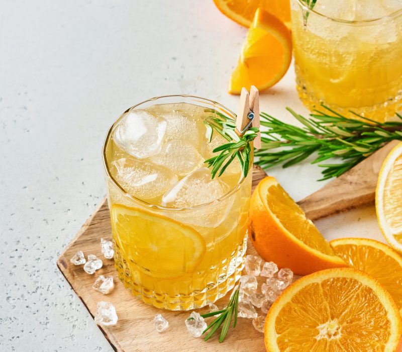 Orange juice or cocktail with rosemary and orange with ice in glass, cold summer lemonade on light gray slate, stone or concrete background. Beach concept with deep sun shadow. Top view.